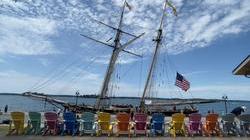 view from the shore, a row of multi-color chairs face a sailboat docked in front of them with a bright blue sky behind it.